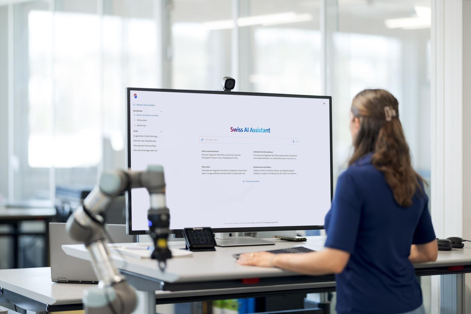 Une personne se tient à un bureau avec un grand écran sur lequel s'affiche l'interface du Swiss AI Assistant. Des équipements de bureau et un bras robotisé sont visibles dans un espace de travail lumineux et moderne.